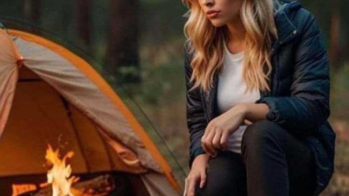 A blond woman in the woods is crouching near the woodfire that's burning INSIDE her tent 